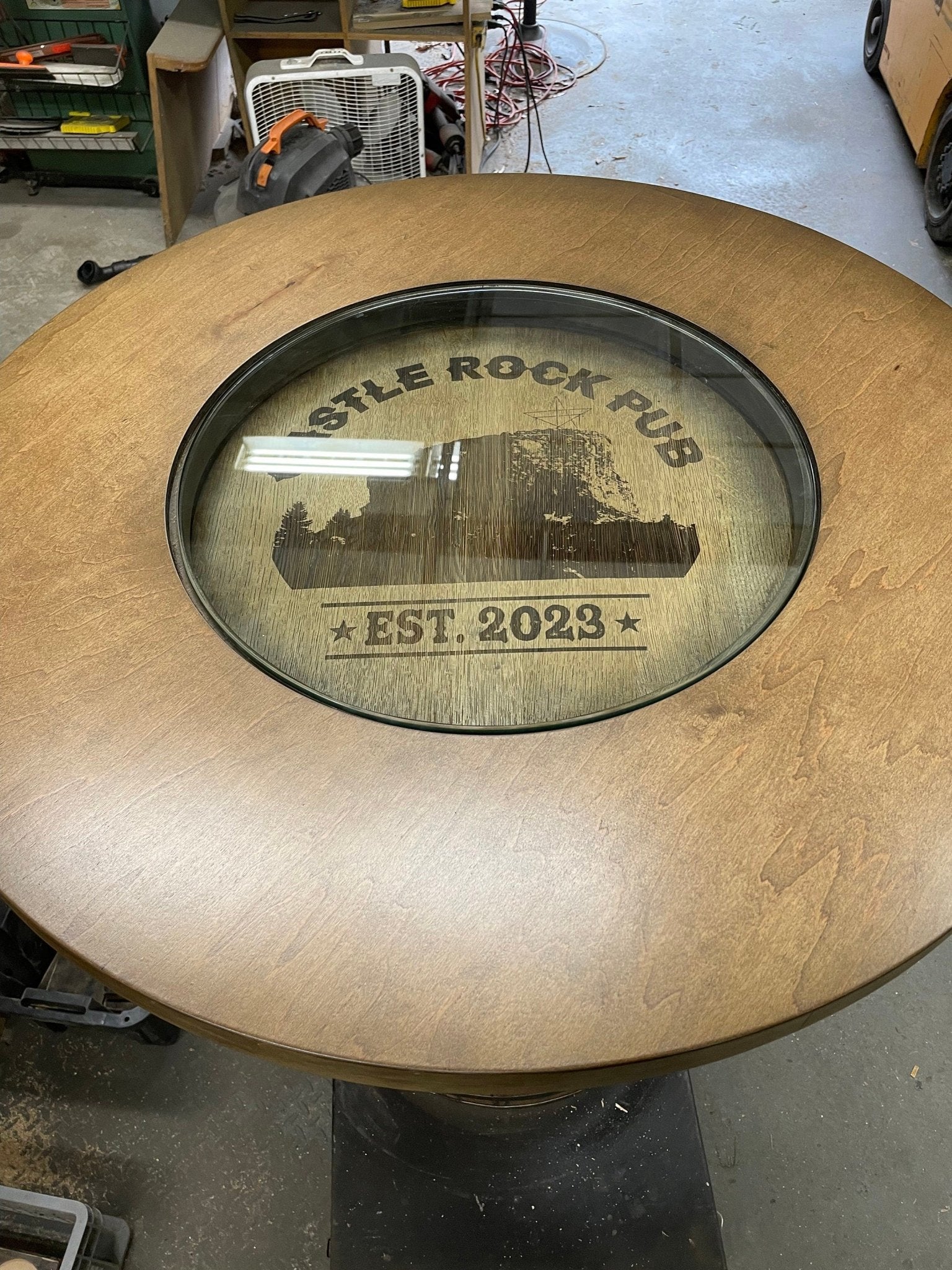 Handcrafted bourbon barrel table with clear glass top insert displaying custom etched pub emblem on reclaimed oak wood head